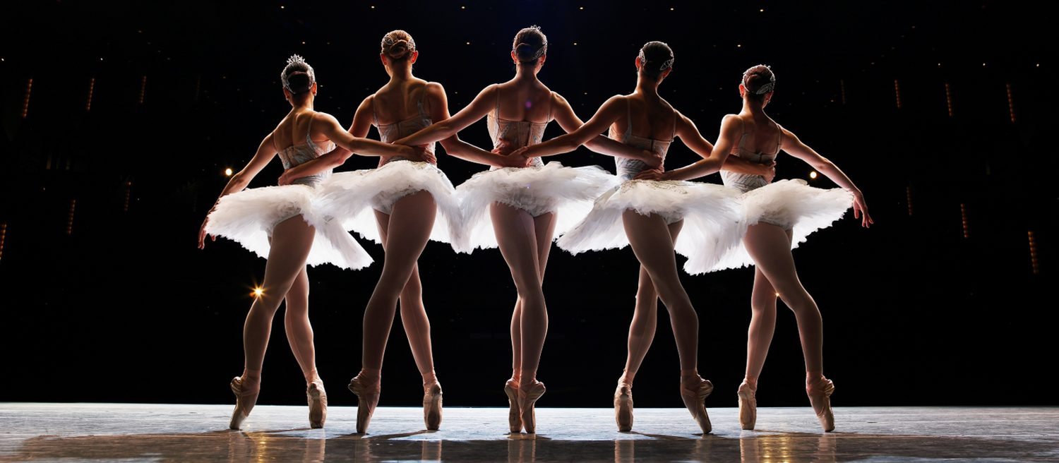 Five dancers on stage, arm in arm, moving as one