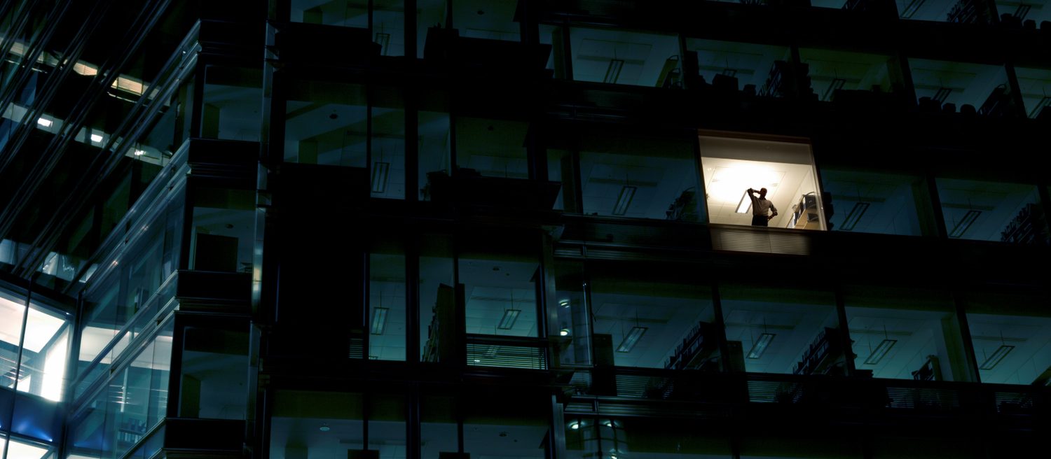 An office building at night, with one lit window and one person in it